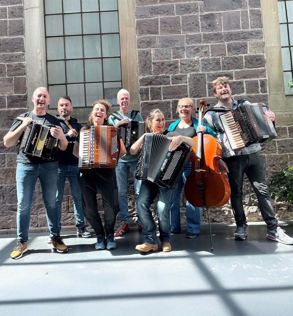 Eine Gruppe von sieben Personen ist zu sehen. Sie stehen vor einer Mauer aus Stein. Fünf von ihnen halten Akkordeons. Eine Person hält ein Chello. Alle lachen.
