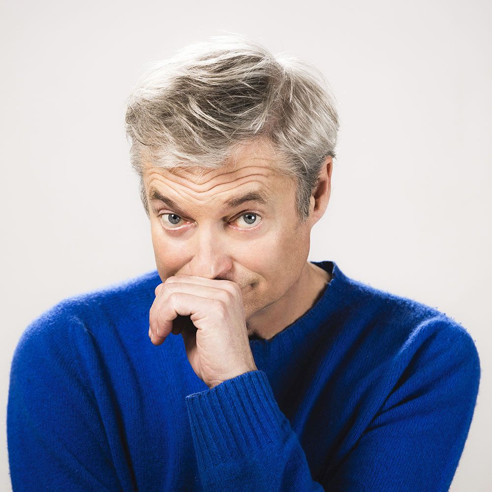 Alfred Dorfer mit grauem Haar in blauem Pullover hält die Hand vor den Mund und blickt direkt in die Kamera