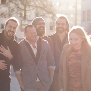 Diie Band John Garner, eine Gruppe von fünf Personen, vier Männer und eine Frau, die im Freien bei Sonnenlicht lachen und entspannt stehen (C) Lisa Seifert