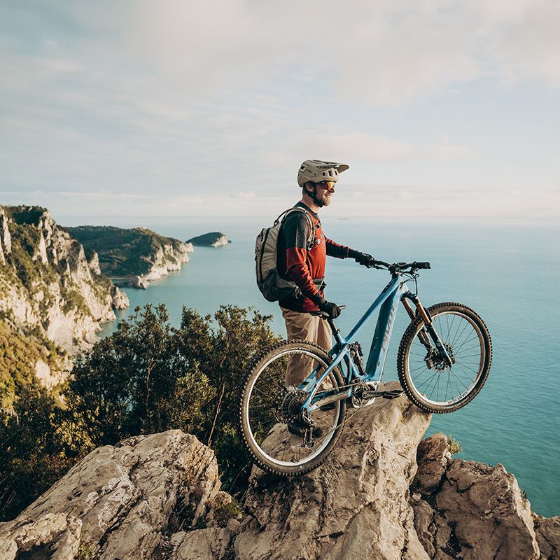 Mann mit Fahrradhelm und Rucksack steht auf Felsen und blickt aufs Meer mit Klippen im Hintergrund © Adrian Greiter