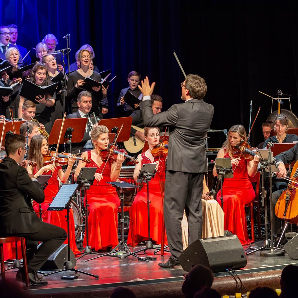 Dirigent vor einem Orchester mit Geigerinnen in roten Kleidern und einem Chor im Hintergrund auf einer Bühne im Parktheater Augsburg (C) Roland Schnell